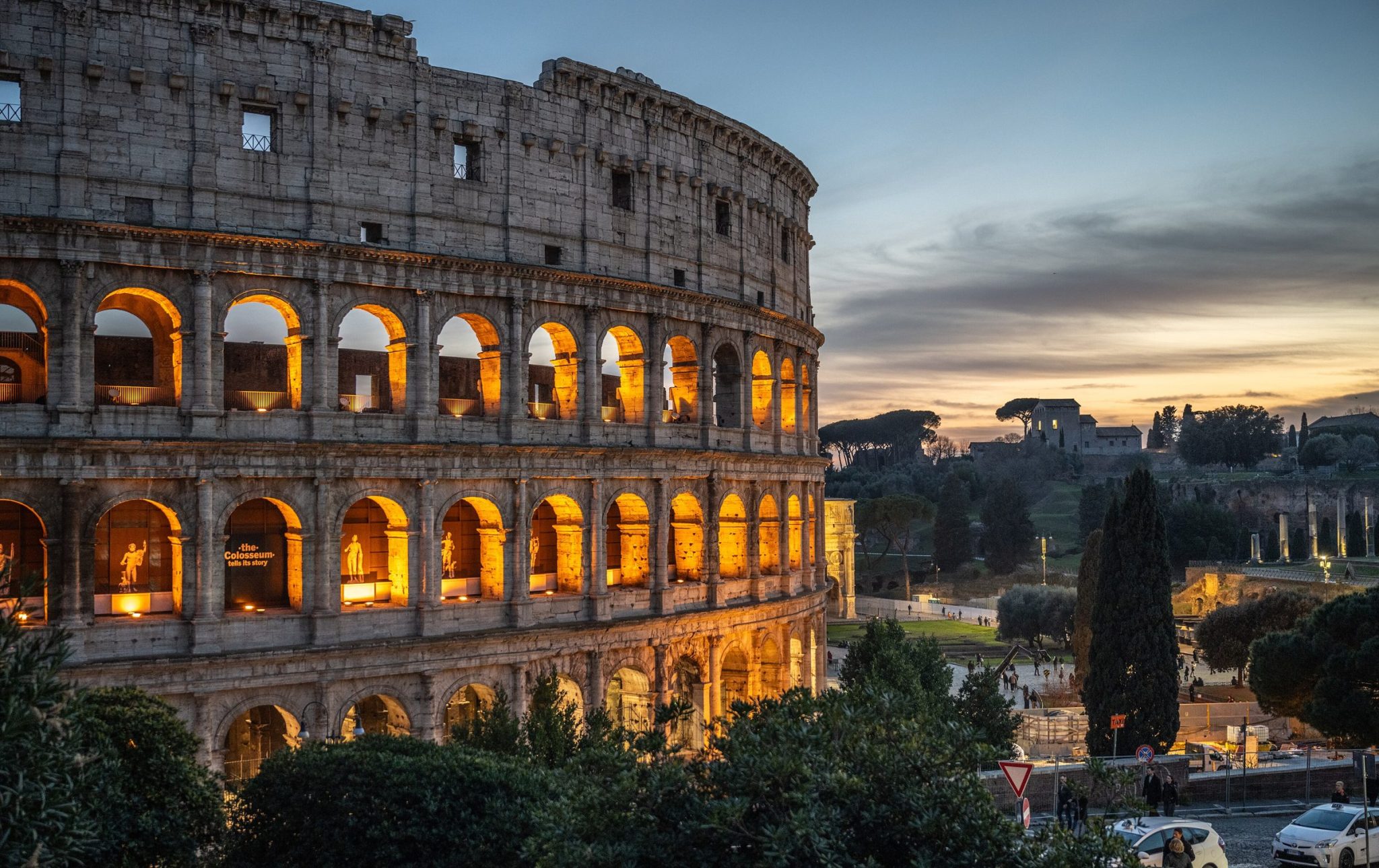 The Colosseum lit from within at dusk, seen through trees